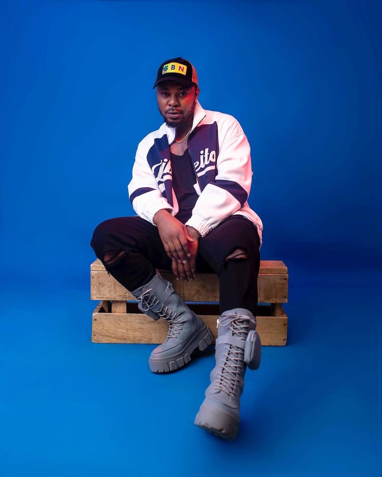 Ogbonna Chinonso (1Notch) - Man sitting on wooden pallet wearing baseball jacket, cap, and gray boots against blue background