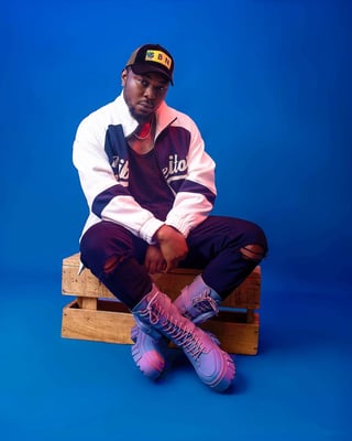 Ogbonna Chinonso 1Notch - Man in varsity jacket and cap sitting on wooden crate against blue background